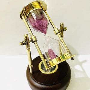 May include: An hourglass with a brass frame and a dark wooden base. The glass bulb contains pink sand. The hourglass is tilted, showing the sand flowing from the top to the bottom. A compass is visible on the base.