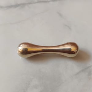 Op de afbeelding: Een gepolijst, goudkleurig metalen object met een afgeronde zandlopervorm. Het gladde oppervlak reflecteert licht, waardoor highlights en schaduwen ontstaan. Het object lijkt een decoratief item of een kleine sculptuur te zijn.