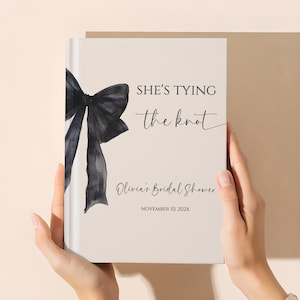May include: A cream-colored bridal shower guest book with a black watercolor bow on the left side. The text "SHE'S TYING the knot" is written in black script. The book also includes the text "Olivia's Bridal Shower" and the date.
