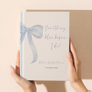 May include: A light beige book with a watercolor-style blue bow and ribbon design. The text on the cover reads "Something blue before I do!" and "Olivia's Bridal Shower" with a date. The book is held by two hands.