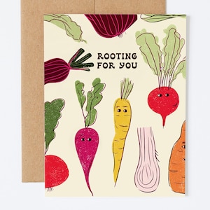 May include: Greeting card with a beige background and illustrations of various vegetables, including beets, carrots, and onions. The text "ROOTING FOR YOU" is displayed in black. The card is paired with a brown envelope.