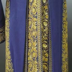 May include: Close-up of a royal blue garment with intricate gold embroidery. The embroidery features floral and scroll patterns along the edges and down the center, creating a rich, textured design. The fabric appears to be a heavy material.
