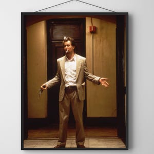 Puede incluir: Impresión enmarcada que presenta a un hombre con traje color beige, camisa blanca y cigarrillo encendido. Está de pie en una puerta, sosteniendo una pistola. La imagen tiene una calidad vintage, tipo película, con tonos cálidos y sensación de drama.