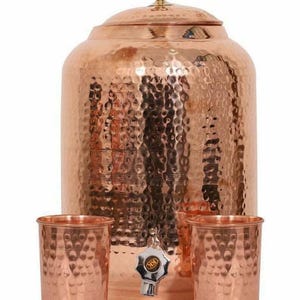 May include: A copper water dispenser with a hammered finish, a tap, a lid, and two matching copper cups. The dispenser has a rounded shape and a golden knob. The cups and dispenser have a textured surface.