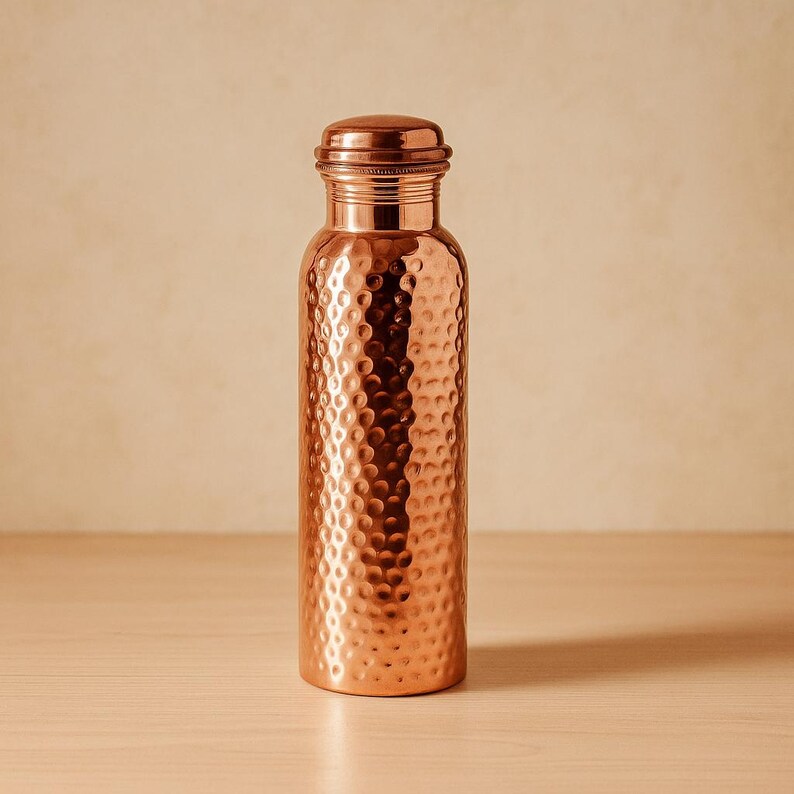 Puede incluir: Una botella de agua de cobre con textura martillada. La botella tiene una base redondeada y una tapa de rosca. El cobre tiene una superficie brillante y reflectante. La botella est&aacute; sobre una superficie de madera clara.