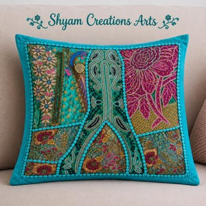Op de afbeelding: Een vierkant sierkussen met een patchworkontwerp van turquoise, magenta en gouden stoffen panelen met ingewikkeld borduurwerk, kralenwerk en bloemmotieven. De tekst Shyam Creations Arts staat in een turquoise scriptlettertype boven het kussen.