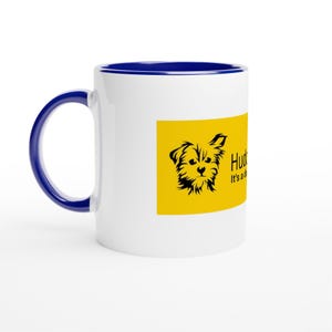 Könnte beinhalten: Weißer Keramikbecher mit blauem Innenraum und Griff. Ein gelbes Rechteck auf dem Becher zeigt eine schwarze Illustration eines Hundes und den Text "Hud... It's a..." in Schwarz. Der Becher ist für Getränke konzipiert.