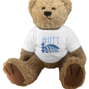Könnte beinhalten: Ein brauner Teddybär, der ein weißes T-Shirt mit dem Text "#UTT" in Hellblau trägt. Das Shirt zeigt auch eine Grafik eines Stadions und zwei blaue Sterne. Der Bär hat dunkelbraune Pfoten und eine schwarze Nase.