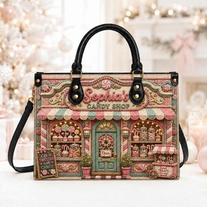 May include: A structured handbag featuring a detailed graphic of a candy shop storefront labeled Sophia's Candy Shop. The design includes a striped awning, a central door, display windows filled with sweets, and a sign reading Life is Sweeter with Candy.