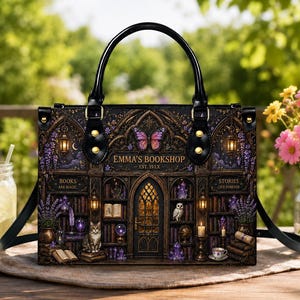 May include: A black handbag featuring a printed graphic of a bookstore interior titled EMMA'S BOOKSHOP EST. 19XX. The design includes bookshelves, a central door, a cat, an owl, crystals, and a butterfly. It has black handles and gold hardware.