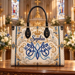 Puede incluir: Un bolso tote estructurado en color crema con borde acolchado, con bordados de filigrana dorada y azul, un icono de corona central y el nombre Sophia en letra cursiva. Tiene dos asas de cuero negro con herrajes en tono dorado y remaches.