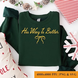 May include: A dark green sweatshirt with the text "His Way Is Better" in yellow script, along with a yellow bow graphic. The sweatshirt is folded neatly, with a Christmas theme. The image also includes a gift box and a snowflake.
