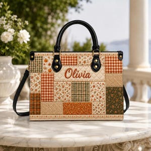 Könnte beinhalten: Eine rechteckige Handtasche mit gestepptem Patchwork-Design in herbstlichen Farben wie Orange, Grün und Creme. Die Tasche hat schwarze Ledergriffe und einen Schulterriemen, wobei der Name Olivia in brauner Schreibschrift vorne aufgestickt ist.