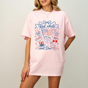 May include: A light pink t-shirt with a patriotic design. The design includes the text "Red, white, and Due" with illustrations of an American flag, fireworks, baby feet, a pacifier, a hot dog, cherries, and a baby bottle.