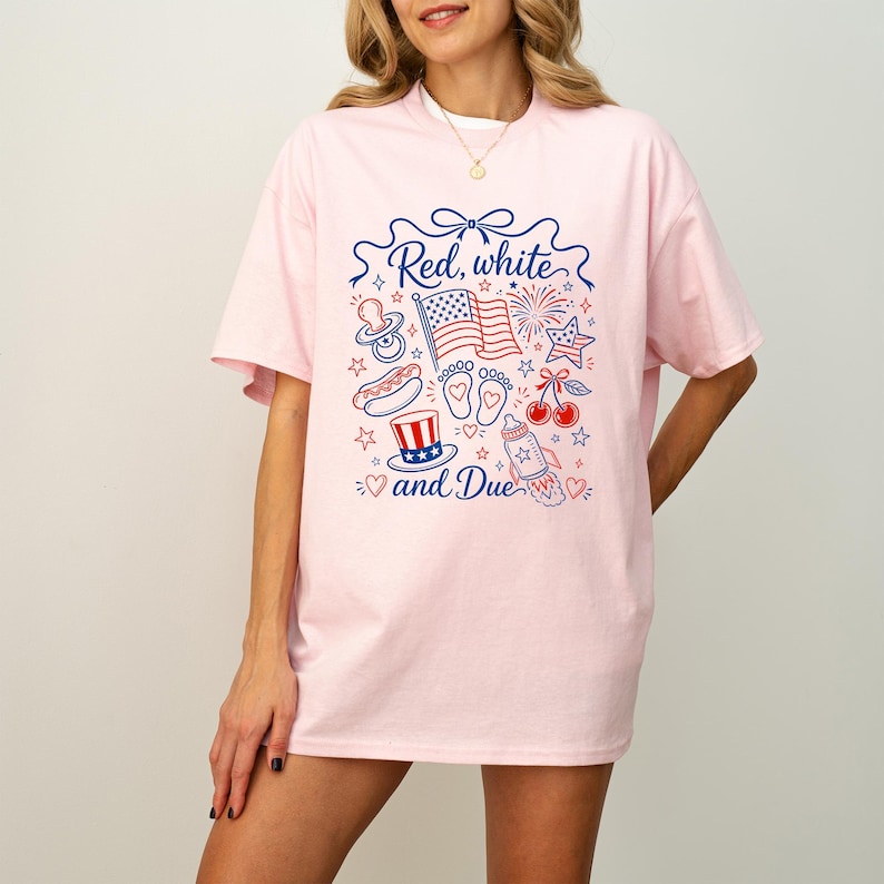 May include: A light pink t-shirt with a patriotic design. The design includes the text "Red, white, and Due" with illustrations of an American flag, fireworks, baby feet, a pacifier, a hot dog, cherries, and a baby bottle.