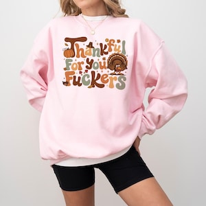 Op de afbeelding: Een lichtroze sweatshirt met de tekst "Thankful For You Fuckers" in bruin, oranje en groen. De afbeelding bevat een pompoen en een kalkoen. De sweatshirt heeft een ronde hals.