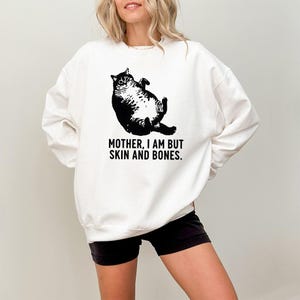 May include: White sweatshirt featuring a black illustration of a cat lying on its back. The text "MOTHER, I AM BUT SKIN AND BONES" is printed below the cat. The sweatshirt is a casual style.
