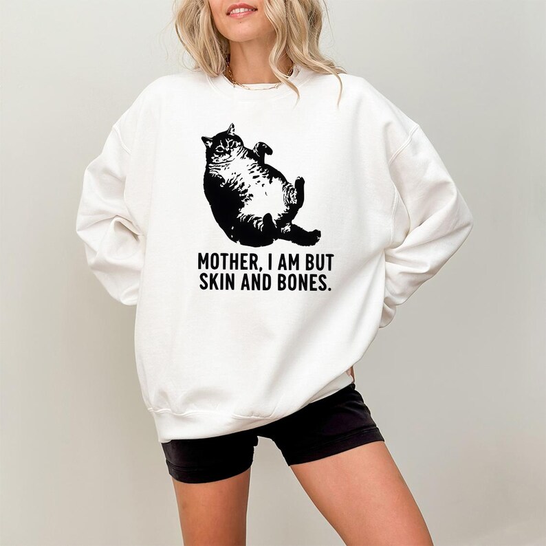 May include: White sweatshirt featuring a black illustration of a cat lying on its back. The text "MOTHER, I AM BUT SKIN AND BONES" is printed below the cat. The sweatshirt is a casual style.