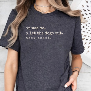 May include: Dark gray t-shirt with the text "it was me. i let the dogs out. they asked." in white. The shirt has short sleeves and a crew neck. The fabric appears to be a soft, comfortable material.