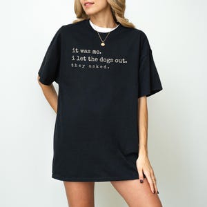 May include: Black t-shirt with the text "it was me. i let the dogs out. they asked." in a white, sans-serif font. The shirt has short sleeves and a crew neck. The model is wearing a white undershirt and a gold necklace.