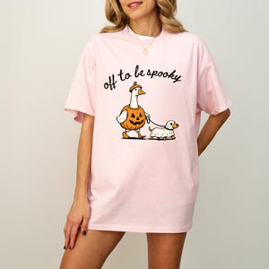 May include: A light pink t-shirt featuring a graphic of a goose wearing a pumpkin costume and walking a small dog dressed as a ghost on a leash. The text above the illustration reads "off to be spooky" in a black, cursive, handwritten-style font.