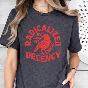 May include: Dark gray t-shirt with a red graphic. The graphic features a bird holding a branch, surrounded by the words "RADICALIZED BY BASIC DECENCY" in a circular design. The shirt has short sleeves.
