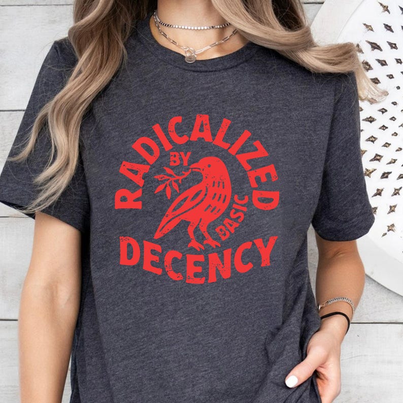 May include: Dark gray t-shirt with a red graphic. The graphic features a bird holding a branch, surrounded by the words "RADICALIZED BY BASIC DECENCY" in a circular design. The shirt has short sleeves.