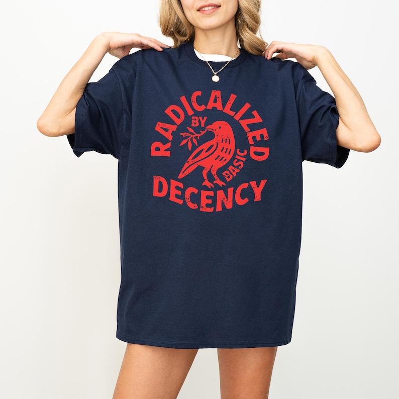 May include: Navy blue t-shirt with a red graphic that reads "RADICALIZED BY BASIC DECENCY" around a bird holding a sprig. The shirt has short sleeves and a crew neck.