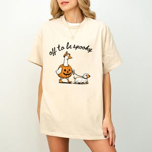 May include: A beige short-sleeve t-shirt featuring a graphic of a goose wearing a pumpkin costume and a small dog wearing a ghost costume. The goose is holding a leash attached to the dog. The text "off to be spooky" is printed in black above.