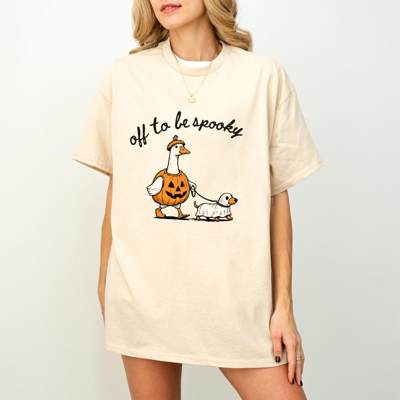 May include: A beige short-sleeve t-shirt featuring a graphic of a goose wearing a pumpkin costume and a small dog wearing a ghost costume. The goose is holding a leash attached to the dog. The text "off to be spooky" is printed in black above.
