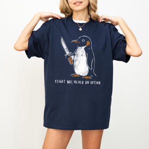 May include: Navy blue t-shirt featuring a cartoon penguin holding a knife. The penguin has a white belly and is outlined in black. The text "FLIGHT WAS NEVER AN OPTION" is printed below the penguin in white.