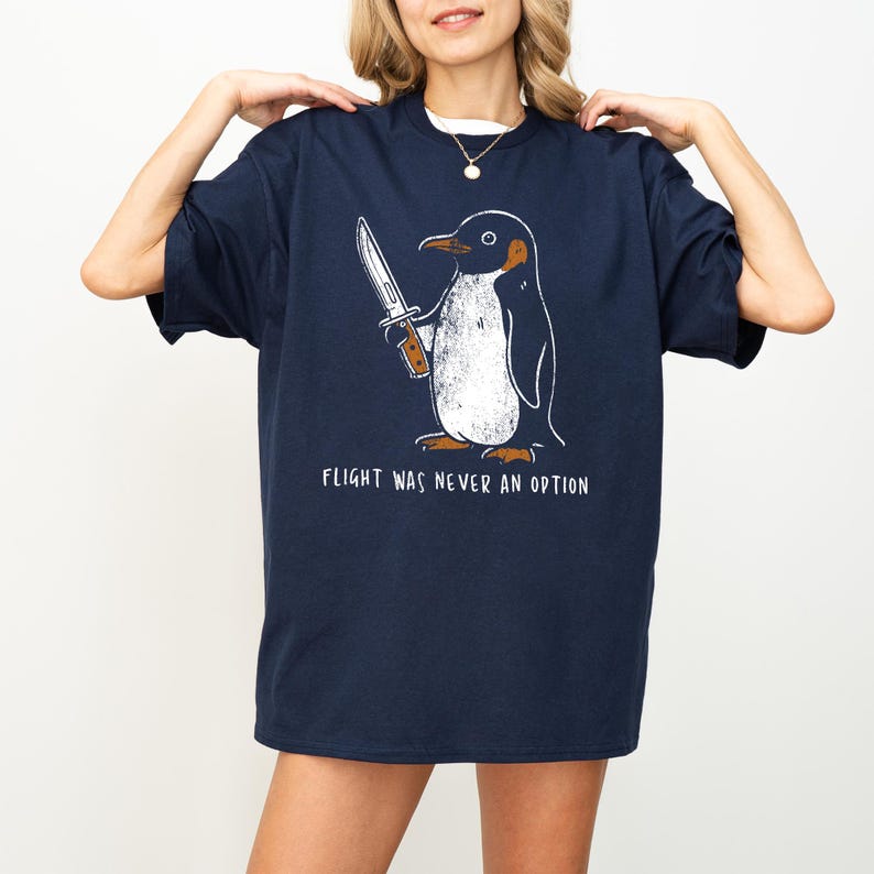 May include: Navy blue t-shirt featuring a cartoon penguin holding a knife. The penguin has a white belly and is outlined in black. The text "FLIGHT WAS NEVER AN OPTION" is printed below the penguin in white.
