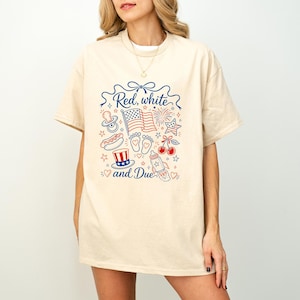 May include: Cream-colored t-shirt with a patriotic design in red and blue. The design includes an American flag, fireworks, a baby bottle, and the text "Red, white and Due".