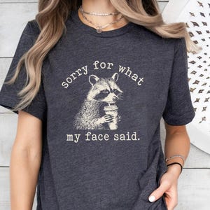 May include: Dark gray t-shirt featuring a raccoon illustration holding a beverage. The text reads "sorry for what my face said." The raccoon is depicted in black and white, with the text in white. The shirt is a casual style.