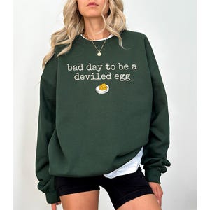 May include: Dark green sweatshirt with the text "bad day to be a deviled egg" in white, along with a small graphic of a deviled egg. The sweatshirt is a casual style.
