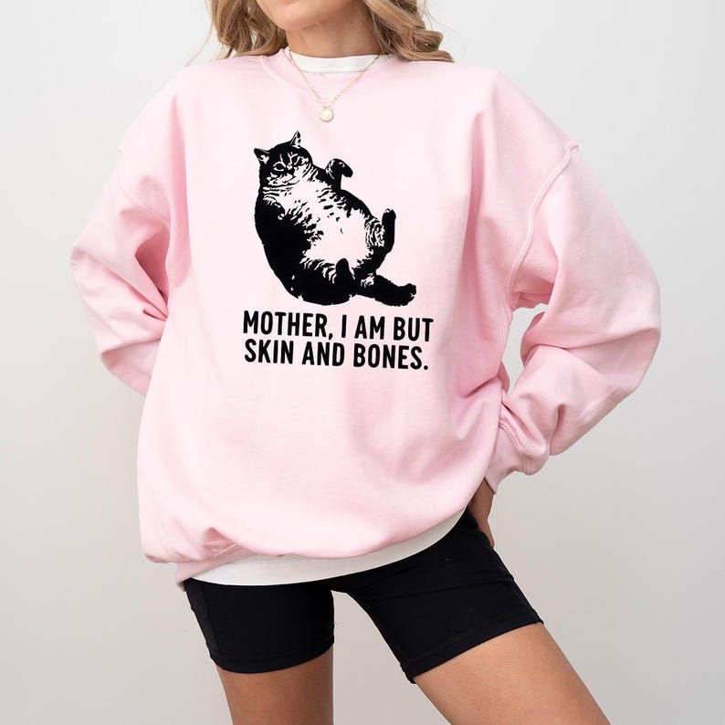 May include: A light pink sweatshirt with a black graphic of a cat lying on its back. The text "MOTHER, I AM BUT SKIN AND BONES." is printed below the cat. The sweatshirt has long sleeves and a crew neck.