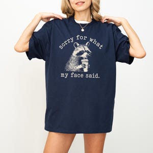 May include: Navy blue t-shirt with a raccoon graphic holding a beverage. The text on the shirt reads "sorry for what my face said." The raccoon illustration is in black and white.