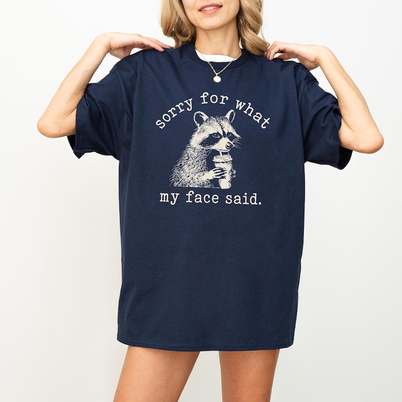 May include: Navy blue t-shirt with a raccoon graphic holding a beverage. The text on the shirt reads "sorry for what my face said." The raccoon illustration is in black and white.