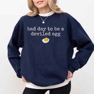 May include: Navy blue sweatshirt with the phrase "bad day to be a deviled egg" in white, along with a small graphic of a deviled egg. The sweatshirt has a crew neck and long sleeves.