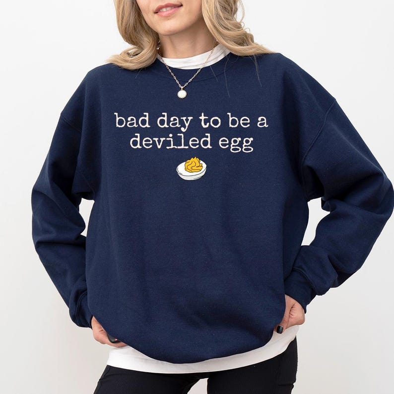 May include: Navy blue sweatshirt with the phrase "bad day to be a deviled egg" in white, along with a small graphic of a deviled egg. The sweatshirt has a crew neck and long sleeves.
