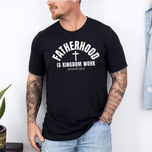 Pu&ograve; includere: T-shirt nera girocollo con testo bianco: "FATHERHOOD IS KINGDOM WORK", un simbolo a croce e "proverbi 22:6". La maglietta &egrave; indossata da una persona con tatuaggi sulle braccia.
