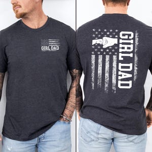K&ouml;nnte beinhalten: Dunkelgraues T-Shirt mit einem verwitterten amerikanischen Flaggen-Design. Auf der Vorderseite steht der Text "GIRL DAD" unter der Flagge. Auf der R&uuml;ckseite des Shirts ist eine Faust-Grafik in einem Flaggen-Design mit vertikalem "GIRL DAD" abgebildet.