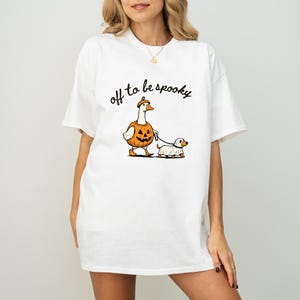 May include: A white short-sleeved t-shirt featuring a graphic of a goose wearing a pumpkin costume and a dog wearing a ghost costume on a leash. The text "off to be spooky" is printed in black cursive script above the illustrated characters.
