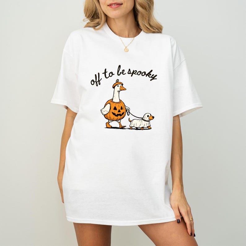 May include: A white short-sleeved t-shirt featuring a graphic of a goose wearing a pumpkin costume and a dog wearing a ghost costume on a leash. The text "off to be spooky" is printed in black cursive script above the illustrated characters.