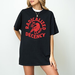 May include: Black t-shirt with a red graphic that reads "RADICALIZED by BASIC DECENCY" around a bird holding a sprig. The shirt has short sleeves and a crew neck, and is worn over a white undershirt.