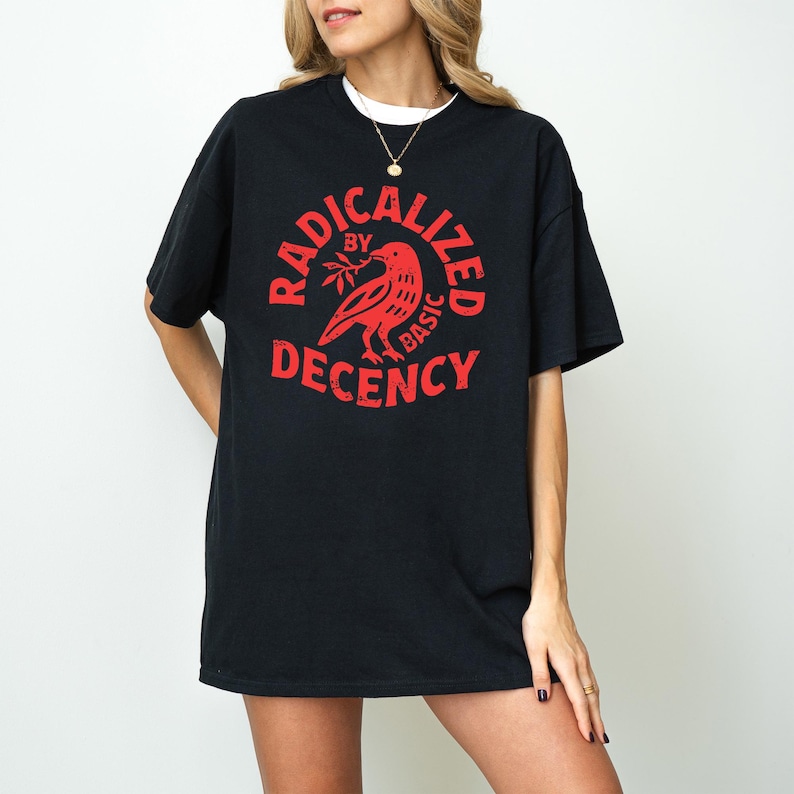 May include: Black t-shirt with a red graphic that reads "RADICALIZED by BASIC DECENCY" around a bird holding a sprig. The shirt has short sleeves and a crew neck, and is worn over a white undershirt.