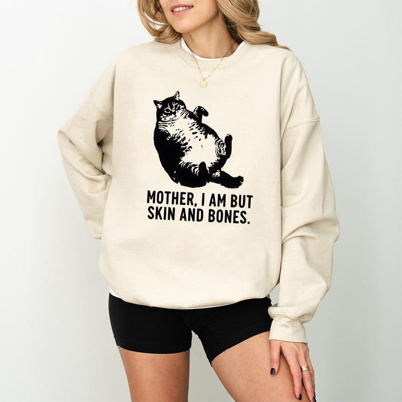 May include: Cream-colored sweatshirt featuring a black graphic of a cat lying on its back. The text "MOTHER, I AM BUT SKIN AND BONES" is printed below the cat. The person in the image is wearing black shorts.