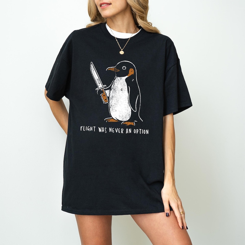 May include: Black t-shirt featuring a cartoon penguin holding a knife. The penguin is in black, white, and orange. The text "FLIGHT WAS NEVER AN OPTION" is printed below the penguin in white.