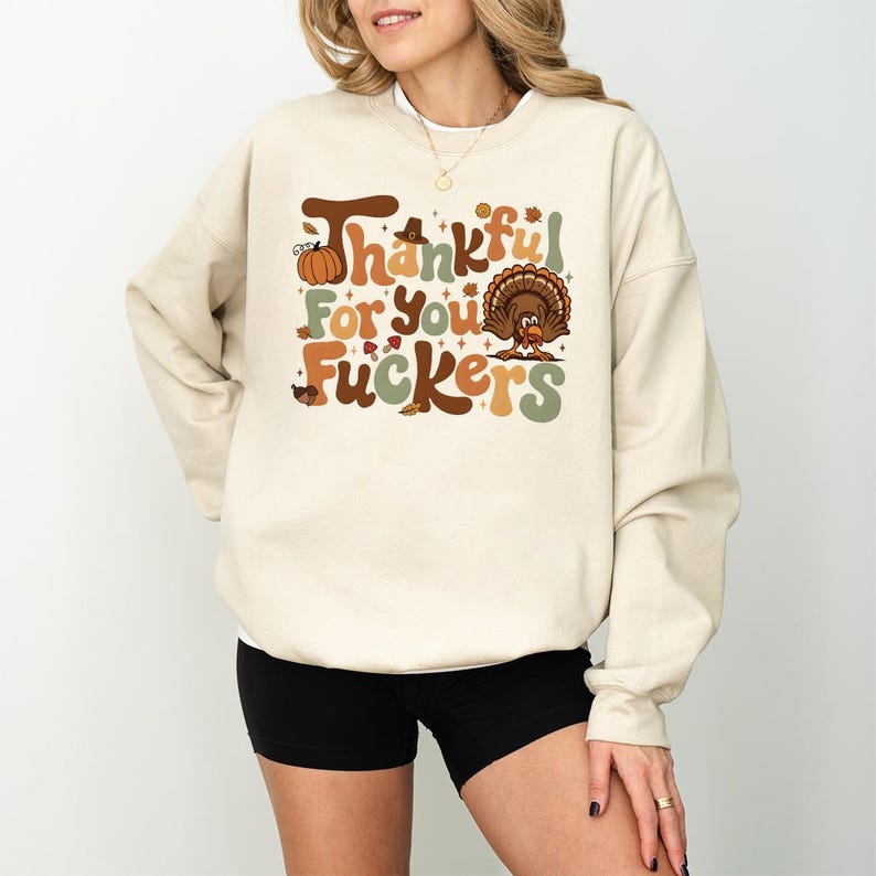 Op de afbeelding: Beige sweatshirt met de tekst "Thankful For You Fuckers" in bruin, oranje en groen. Het ontwerp bevat een pompoen, een pelgrimshoed en een kalkoen. Het sweatshirt wordt gedragen met een zwarte short.