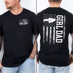 K&ouml;nnte beinhalten: Schwarzes T-Shirt mit einem verblassten amerikanischen Flaggen-Design. Auf der Vorderseite steht "GIRL DAD" unter der Flagge. Auf der R&uuml;ckseite ist eine Faust im Flaggen-Design abgebildet, mit vertikal gedrucktem "GIRL DAD".